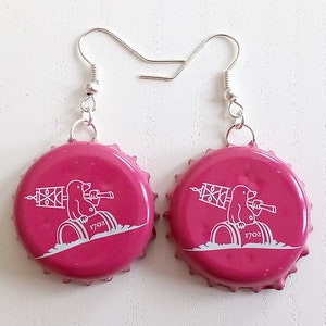 Puede incluir: Un par de pendientes de tapón de botella rosa con un diseño blanco de un topo sosteniendo una bandera y sentado en un barril. El texto "1702" está impreso en el barril.