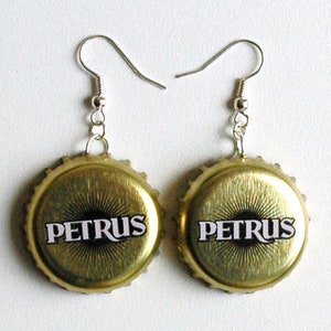Peut inclure: Une paire de boucles d'oreilles en forme de capsule de bouteille dorée avec des crochets argentés. Les capsules de bouteille ont le mot "PETRUS" imprimé en blanc.