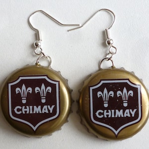 Puede incluir: Un par de pendientes de tapón de botella dorado con un diseño de escudo marrón con dos flores de lis blancas y la palabra "CHIMAY" en letras blancas.