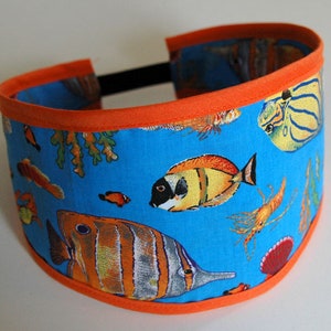 Peut inclure: Un bandeau avec une bordure orange et un fond bleu représentant des poissons et des coraux colorés. Le bandeau est en tissu et possède un élastique extensible.
