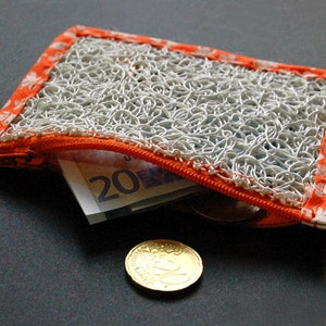 Könnte beinhalten: Eine kleine orangefarbene und weiße Reißverschlusstasche mit einem weißen Netzfutter. Die Tasche ist offen und enthält einen Zwanzigeuro-Schein und eine Münze.
