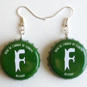 Puede incluir: Un par de pendientes de tapón de botella verde con ganchos plateados. Los tapones de botella tienen el texto "Biere de l'Abbaye de Floreffe" y una "F" blanca.