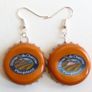 Könnte beinhalten: Ein Paar handgefertigte Ohrringe mit orangefarbenen Kronkorken und einem blau-weißen Etikett. Auf dem Etikett steht "Queue de Charrue Ploegsteert". Die Ohrringe haben silberfarbene Ohrhaken.