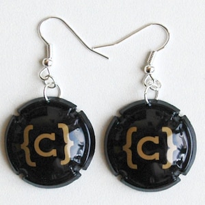 Puede incluir: Un par de pendientes negros y dorados hechos con tapones de botellas de champán. Los pendientes presentan la letra "C" en dorado con llaves alrededor.