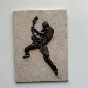 May include: A rock art sculpture of a person playing a guitar. The sculpture is made of small, dark brown rocks and is mounted on a beige tile.