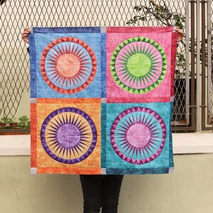 May include: A colourful fabric quilt with four squares. Each square features a sunburst design in a different colour: red, green, purple, and blue. The quilt is made of a soft, textured fabric.