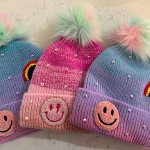 May include: Three colorful knit hats with pom-poms. Each hat features a different embroidered design: a rainbow, a smiley face, and a smiley face with a lightning bolt. The hats are decorated with iridescent sequins.