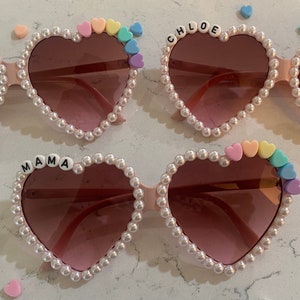 Op de afbeelding: Drie paar hartvormige zonnebrillen met parelversiering. Elk paar heeft een andere naam in zwarte letters op het montuur: "EMMA", "CHLOE" en "MAMA". Elk paar heeft ook een regenboog van pastelkleurige hartparels aan &eacute;&eacute;n kant.