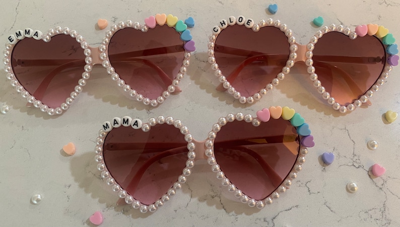 Op de afbeelding: Drie paar hartvormige zonnebrillen met parelversiering. Elk paar heeft een andere naam in zwarte letters op het montuur: "EMMA", "CHLOE" en "MAMA". Elk paar heeft ook een regenboog van pastelkleurige hartparels aan &eacute;&eacute;n kant.