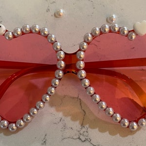 Op de afbeelding: Hartvormige zonnebril met een roze montuur en witte parel accenten. De zonnebril heeft de naam "Alice" in witte letters op de linkerlens.