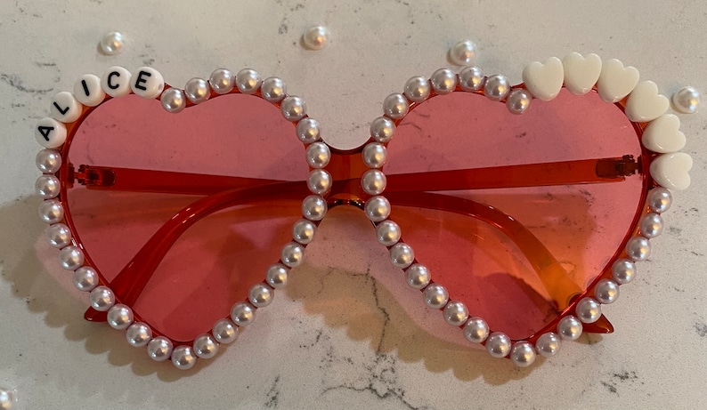 Op de afbeelding: Hartvormige zonnebril met een roze montuur en witte parel accenten. De zonnebril heeft de naam "Alice" in witte letters op de linkerlens.