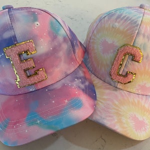 May include: Two tie-dye baseball caps with pink and blue colors and a white background. The caps have a large pink letter on the front, one says "E" and the other says "C".