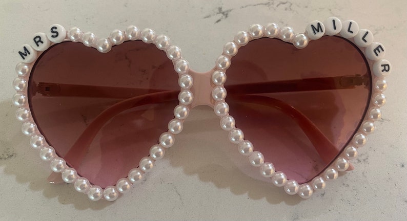 Op de afbeelding: Roze hartvormige zonnebril met witte parel accenten. De zonnebril heeft de woorden "MRS" aan de ene kant en "MILLER" aan de andere kant.
