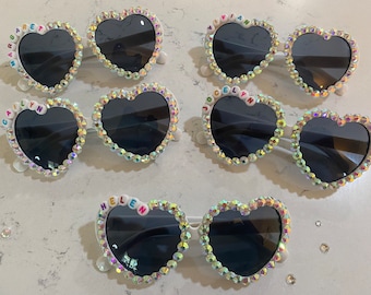 May include: Five pairs of white heart-shaped sunglasses with dark lenses. Each pair is embellished with iridescent rhinestones around the frames. Some frames feature colorful letter beads spelling the names Margaret, Vivian, Cailyn, Jocelyn, and Helen.