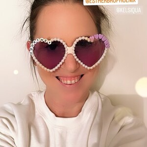 Op de afbeelding: Een vrouw draagt hartvormige zonnebrillen met parel- en paarse accenten. De zonnebril heeft de naam "COURTNEY" op het linker glas. De vrouw draagt een wit sweatshirt met de tekst "COOLER THAN" en de Instagram-handle "@ARIANAMADIX" erop gedrukt. De tekst "Loving my new sunglasses!" staat boven het hoofd van de vrouw, samen met de Instagram-handles "@ESTHERBROPHOENIX" en "@KELSISQUA".