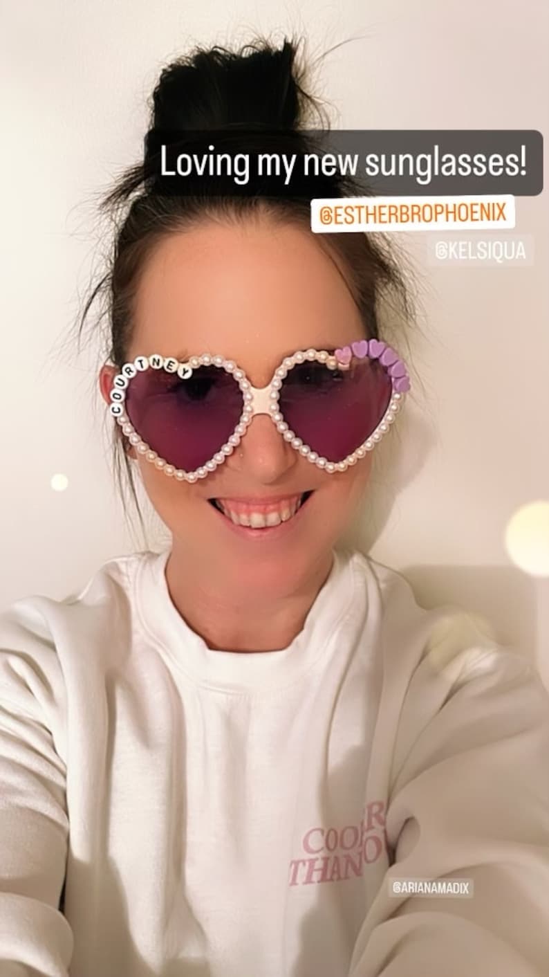 Op de afbeelding: Een vrouw draagt hartvormige zonnebrillen met parel- en paarse accenten. De zonnebril heeft de naam "COURTNEY" op het linker glas. De vrouw draagt een wit sweatshirt met de tekst "COOLER THAN" en de Instagram-handle "@ARIANAMADIX" erop gedrukt. De tekst "Loving my new sunglasses!" staat boven het hoofd van de vrouw, samen met de Instagram-handles "@ESTHERBROPHOENIX" en "@KELSISQUA".