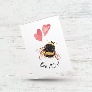 May include: A white card with a watercolour illustration of a bumblebee and two pink hearts. The card says "Bee Mine!"
