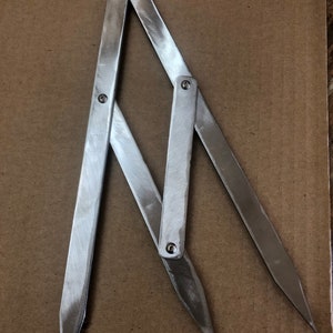 May include: A silver metal compass tool with two arms and a pointed tip on each end. The arms are joined together with a hinge and have a brushed finish.