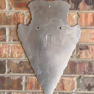 May include: A silver metal arrowhead shaped wall decor with two holes for hanging. The arrowhead is mounted on a brick wall.