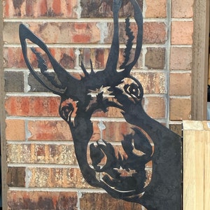 May include: A black metal donkey silhouette wall decor with a whimsical expression. The donkey's head is facing the right side of the image, and the donkey's ears are pointed up. The donkey is mounted on a brick wall.