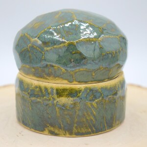 May include: A handcrafted ceramic jar with a lid. The jar is a combination of green and yellow hues, with a textured surface. The lid is rounded, and the base is cylindrical. The jar is likely used for storage or decoration.