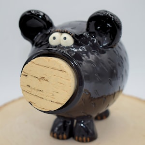 May include: A whimsical black ceramic pig-shaped bottle stopper with a cork. The pig has large, expressive eyes and ears. The stopper is designed to fit into a bottle opening. The pig is on a wooden surface.