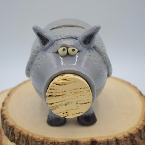May include: A whimsical, gray ceramic pig-shaped bottle stopper with a cork. The pig has large, expressive eyes and ears. The stopper is resting on a wooden surface. The pig has a light gray body and dark gray accents.