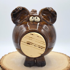 May include: A round, glossy brown ceramic piggy bank featuring large, expressive eyes and prominent ears. The front of the pig has a circular opening sealed with a light-colored, textured cork stopper. The figure stands on four small, dark hooves.
