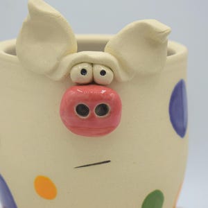 May include: A ceramic pig-themed mug with a cream-colored base and colorful polka dots. The mug features a pig face with a pink snout, black eyes, and large ears. The mug is approximately 4 inches tall.