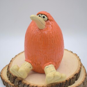 May include: A whimsical orange ceramic bird figurine with a textured surface, sitting on a wooden slice. The bird has a light beige beak and feet, and is posed in a seated position. The figurine is a unique decorative item.