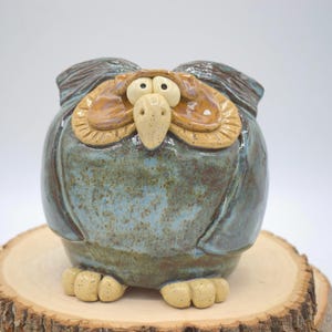 May include: A handcrafted ceramic owl figurine with a whimsical design. The owl features a blue-green body, tan beak, and large, expressive eyes. The owl is perched on a wooden slice, showcasing its unique, artistic style.