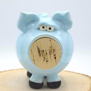 May include: A light blue ceramic piggy bank shaped like a pig with large ears and wide eyes. The center of the pig features a circular tan cork stopper. The pig has two small front legs with dark brown hooves at the base of the round body.