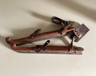 May include: A pair of antique wooden ice skates with leather straps. The skates are brown and have a worn, rustic look.
