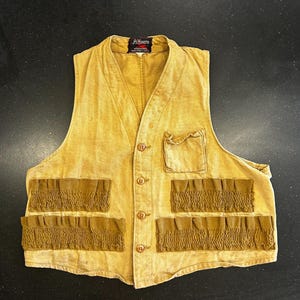 May include: A vintage J.C. Higgins hunting vest in a faded tan color. The vest has a V-neck, button closure, and multiple pockets, including four rows of cartridge holders in a darker tan. The vest has a worn appearance.