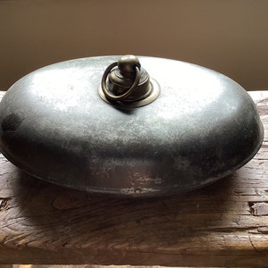 May include: A silver oval-shaped container with a brass lid and a ring handle. The container is sitting on a wooden surface.