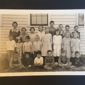 May include: A black and white photo of a group of children, mostly girls, standing in front of a wooden building. The children are all dressed in clothing from the early 1900s. Some of the children are smiling, while others have serious expressions.