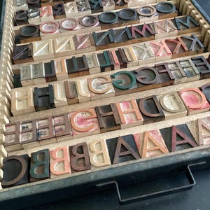 May include: A vintage wooden printing block case filled with letterpress type. The case is made of dark wood and has a metal handle. The type is arranged in rows and columns, and includes letters, numbers, and punctuation marks.
