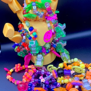 Op de afbeelding: Een verzameling kleurrijke kralenarmbanden en accessoires, waaronder neon groene, roze, oranje en paarse kralen. De armbanden worden getoond op een gele handvormige standaard, wat een levendige en speelse uitstraling creëert.