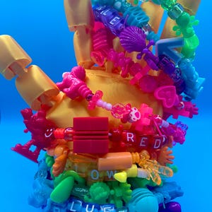 May include: A yellow articulated hand model adorned with colorful beaded bracelets. The bracelets feature various shapes and the words "BLUE", "RED", and "LOW". The background is a solid blue.