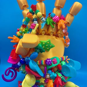 May include: A gold-colored articulated mannequin hand display stand adorned with a dense collection of colorful plastic beads, charms, and miniature objects including stars, hands, caps, and spiral shapes in various bright primary and neon colors.