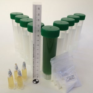 May include: A collection of clear, cylindrical tubes with green caps, some filled with clear liquid, others with green liquid. A ruler with centimeter markings stands next to the tubes. Small vials and a bag of white powder are also present. The text "Algae Research and Supply" is visible.