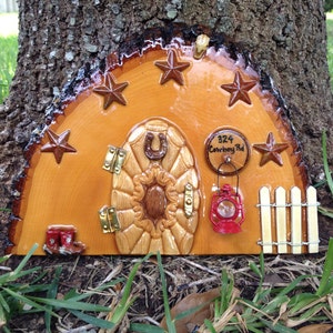 May include: A wooden fairy door with a horseshoe, a carved sunflower design, and a red lantern. The door is painted brown and has a sign that reads "324 Cowboy Rd".