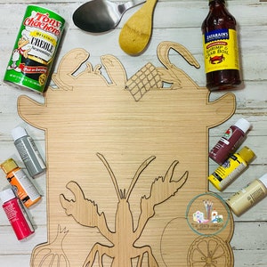 May include: Wooden cutting board with a lobster, crab, lemon, and garlic design. The board is surrounded by paint bottles, a wooden spoon, and a bottle of Zatarain's Shrimp & Crab Boil.