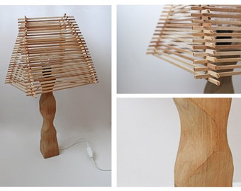 Può includere: Una lampada da tavolo in legno con una base in legno naturale e un paralume intrecciato con sottili strisce di legno. Il paralume è quadrato e contiene una lampadina.