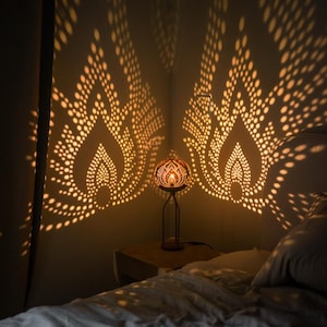 May include: A patterned table lamp, projecting intricate light designs onto the wall. The lamp is on a wooden bedside table next to a bed with white bedding. The warm light creates a cosy atmosphere in the room.