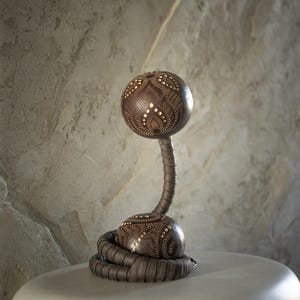 Może przedstawiać: Dekoracyjna lampa stołowa z ciemnobrązowym, kulistym kloszem i podstawą, ozdobiona skomplikowanymi wzorami w kropki. Trzon lampy ma elastyczną, teksturowaną konstrukcję, a podstawa jest otoczona zwiniętym, plecionym elementem. Lampa stoi na białej powierzchni.