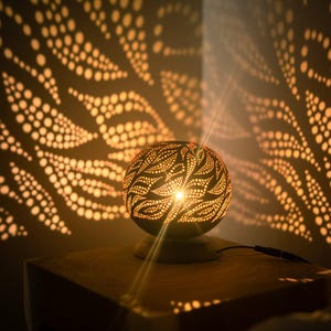 Può includere: Una lampada sferica decorativa con intricati motivi a pois, che proietta ombre a forma di foglia sulla parete. La calda luce emana dall'interno, creando un'atmosfera accogliente. La lampada è posizionata su una superficie di legno.