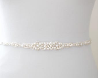 May include: Ivory-colored bridal belt with a floral design. The belt features small pearls and sparkling crystals. The belt is designed to be worn around the waist. The belt is made of fabric and is a delicate accessory.