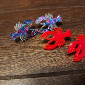 May include: Three lobster-shaped earrings. One pair is red, and the other pair is blue and pink with glitter.