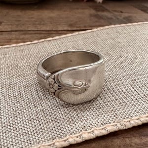 Puede incluir: Un anillo de plata hecho de una cuchara, con detalles florales y de volutas. El anillo tiene una banda ancha y una estética vintage. El anillo se muestra sobre una tela de tono neutro, con un fondo de madera.
